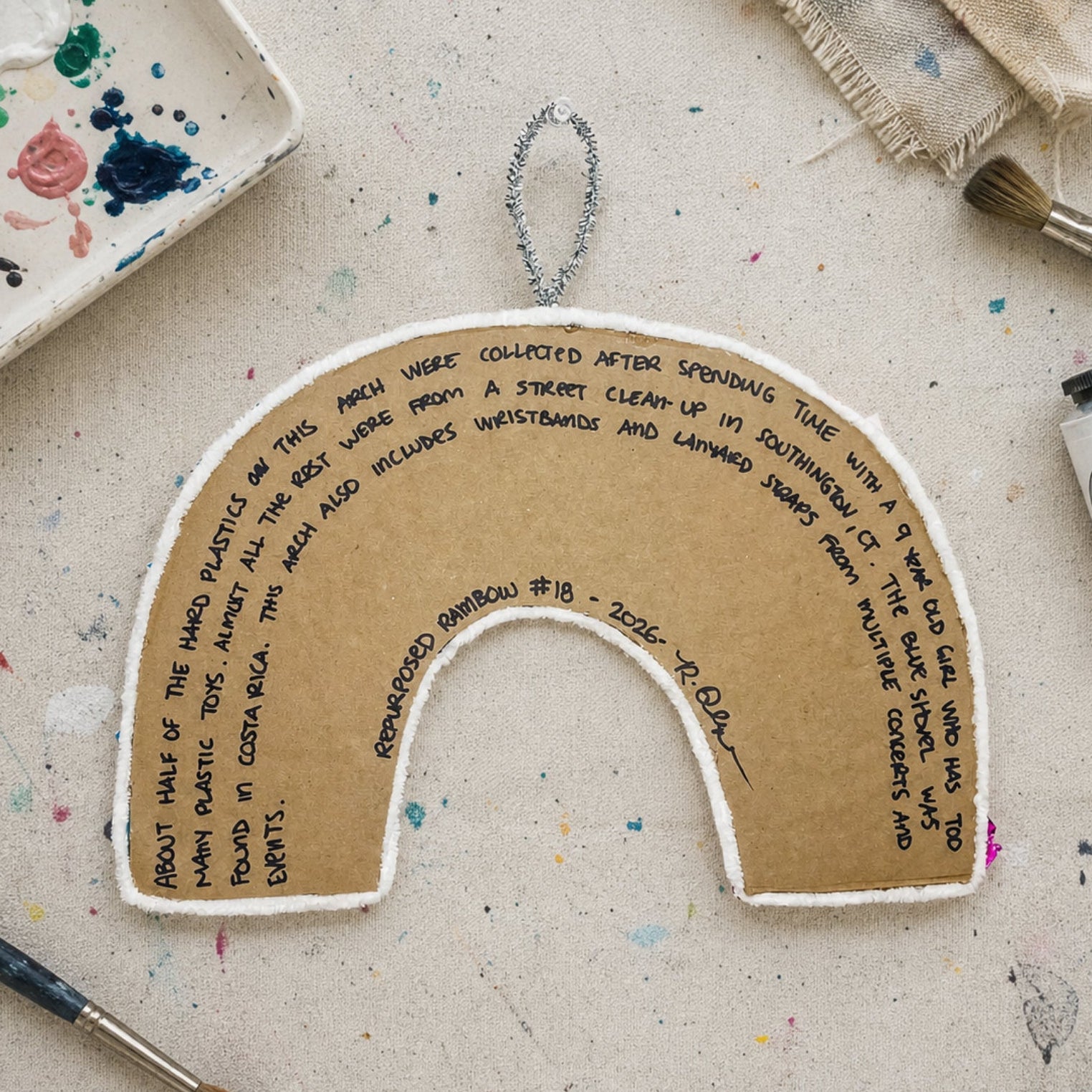 Handmade rainbow-shaped craft item with text on a textured surface with art supplies.